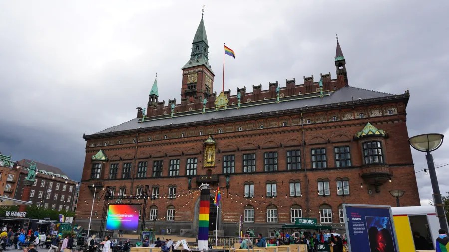 Blick auf das Kopenhagener Rathaus mit Regenbogenflaggen und einem bunten Pride-Stand im Vordergrund, unter einem bewölkten Himmel.