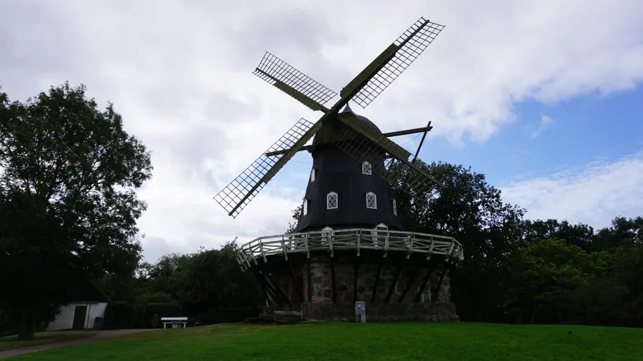 Eine historische Windmühle steht auf einer Wiese, umgeben von Bäumen und einem bewölkten Himmel.