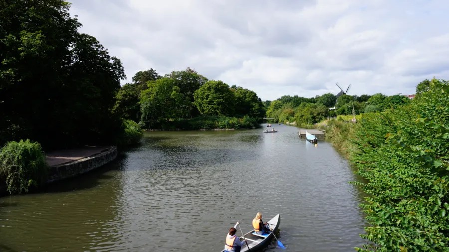 A serene view of a river surrounded by lush greenery, with two people kayaking in the foreground and a small boat in the distance under a cloudy sky.