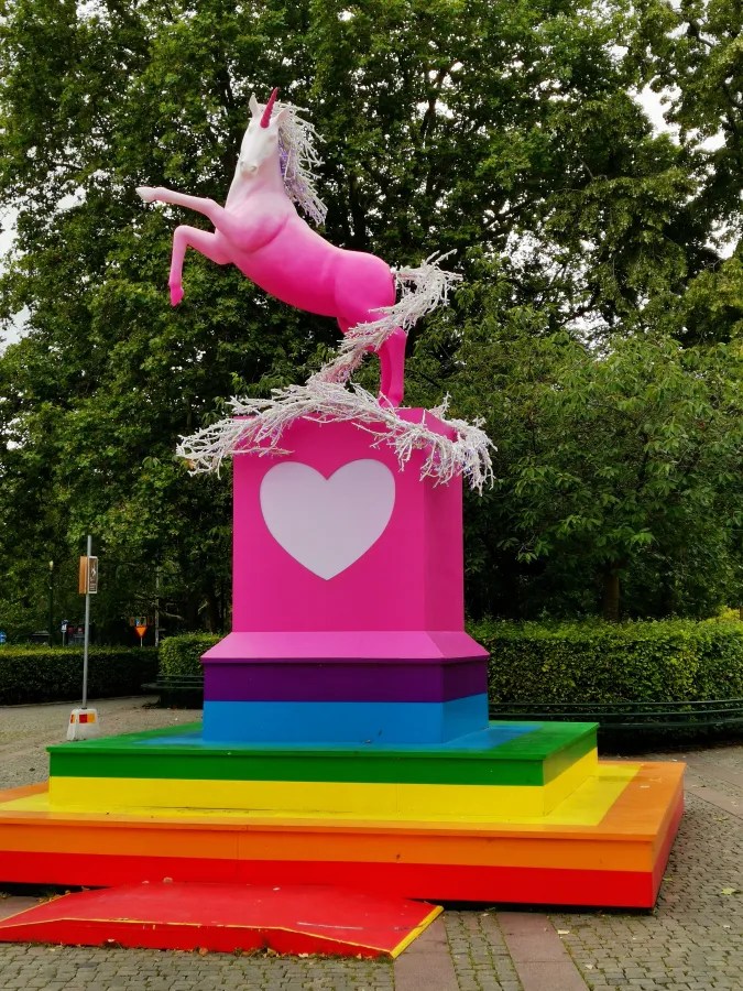 Ein pinkes Einhorn, das auf einem mehrfarbigen Sockel mit einem Herzsymbol steht, umgeben von grünen Bäumen.