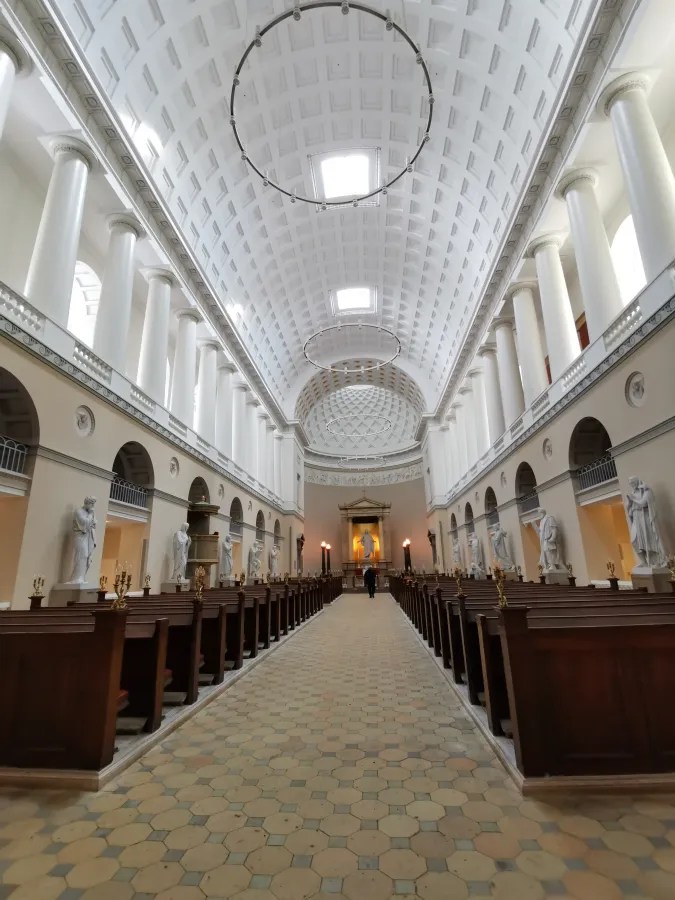 Blick in das Innere einer Kirche mit hohen Säulen, einem gewölbten Deckenbereich und einer langen Mittelachse, die zu einem Altar führt. Mehrere Statuen sind entlang der Wände aufgestellt.
