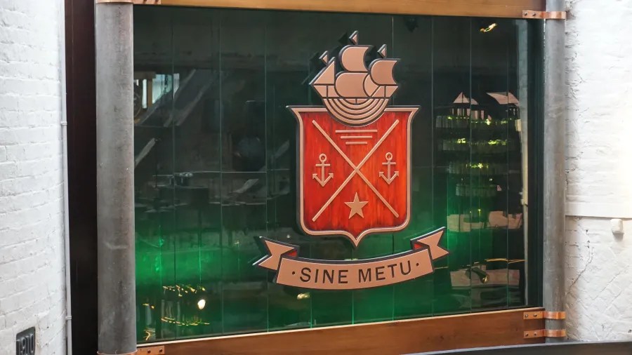 Ein farbenfrohes Wappen mit einem Schiff und Ankern, darunter der Schriftzug 'SINE METU'.