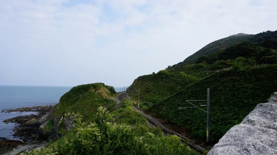 Blick auf die Küste von Irland mit grünen Hügeln und einem Eisenbahngleis, das entlang der Küste verläuft.