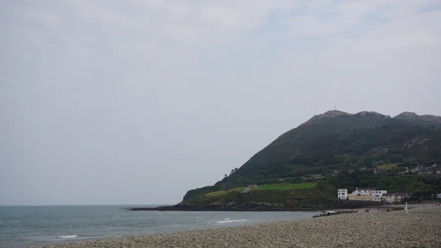 Blick auf einen Strand mit Kieselsteinen und einer sanften Küstenlinie, im Hintergrund ein grüner Hügel, ein bewölkter Himmel und einige Gebäude in der Nähe des Wassers.