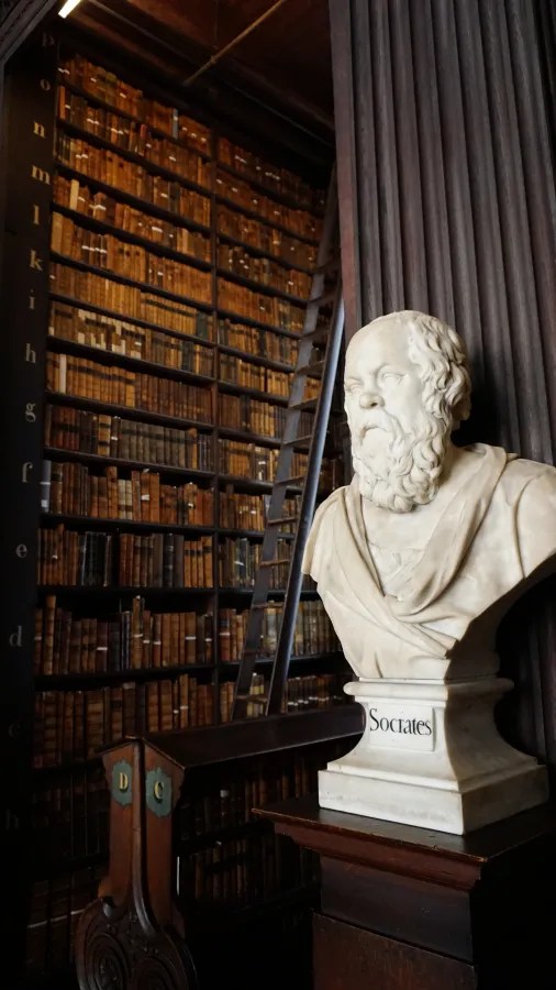 Ein marmorner Büstenkopf von Sokrates steht vor einer hohen Bücherwand in einer historischen Bibliothek. Die Regale sind voller alter Bücher mit Lederbinden, und eine Leiter ist sichtbar, um die oberen Regale zu erreichen.