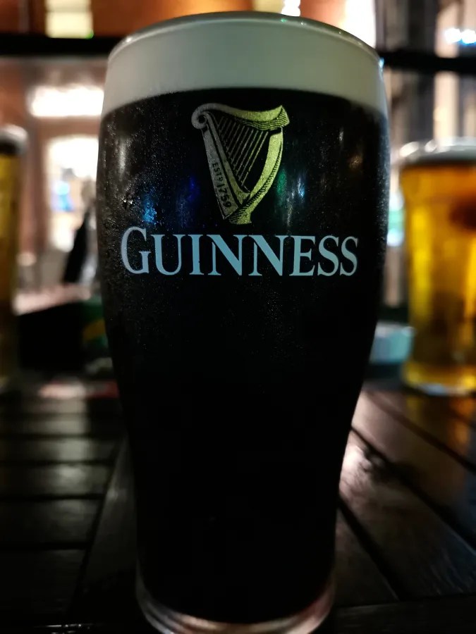 Ein Glas Guinness, ein bekanntes irisches Bier, auf einem Tisch in einem Pub, umgeben von verschwommenen Getränken im Hintergrund.