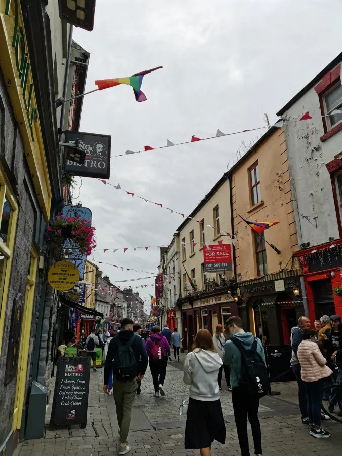 Lebhafte Straße in Galway mit farbenfrohen Geschäften, Menschen und bunten Fähnchen, darunter eine Regenbogenflagge.