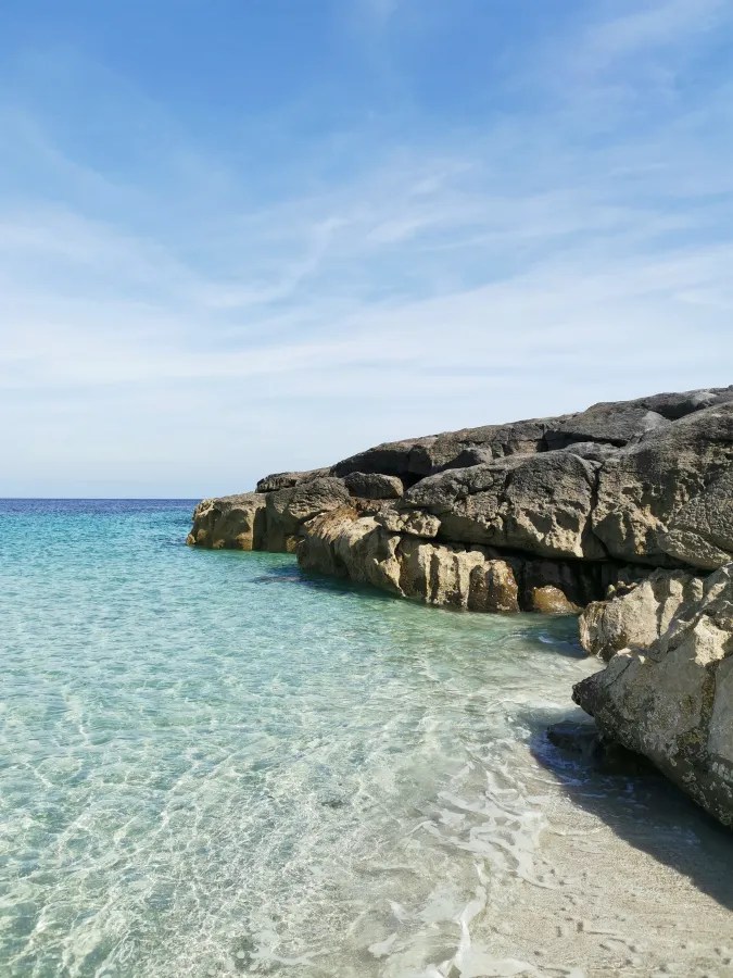 Klarer blauer Himmel über einem ruhigen Strandabschnitt mit transparentem Wasser und großen Felsen, die umspült werden.