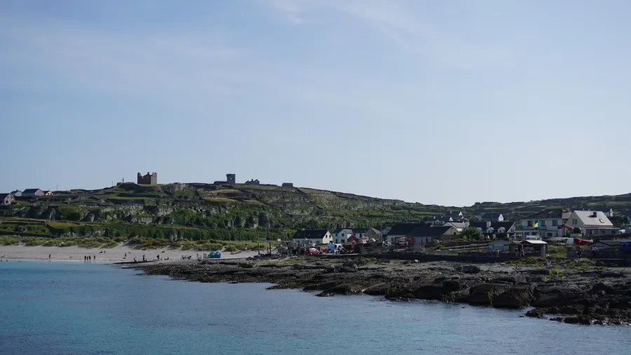 A serene view of Inis Oírr, one of the Aran Islands in Ireland, showing a rocky shoreline, a small village with houses, and grassy hills in the background under a clear blue sky.