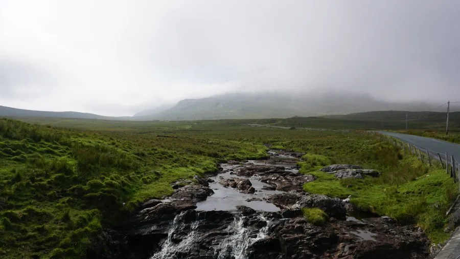 Eine sanfte, bewölkte Landschaft in Connemara mit grünen Wiesen, einer kleinen Wasserstelle und sanften Hügeln im Hintergrund.