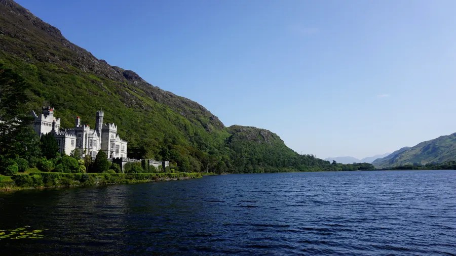 Kylemore Abbey nestled beside a tranquil lake, surrounded by lush green hills under a clear blue sky.