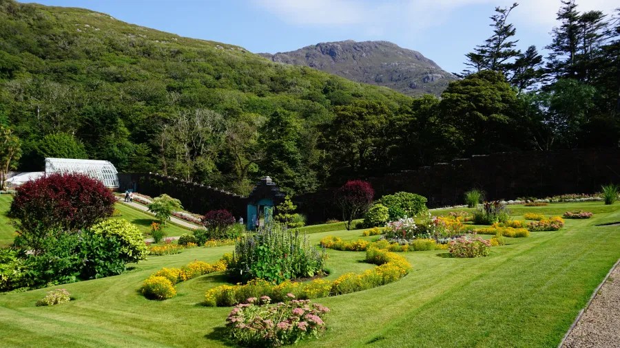 Ein gepflegter Garten mit bunten Blumen und grünen Rasenflächen, umgeben von sanften Hügeln und Bäumen im Hintergrund.