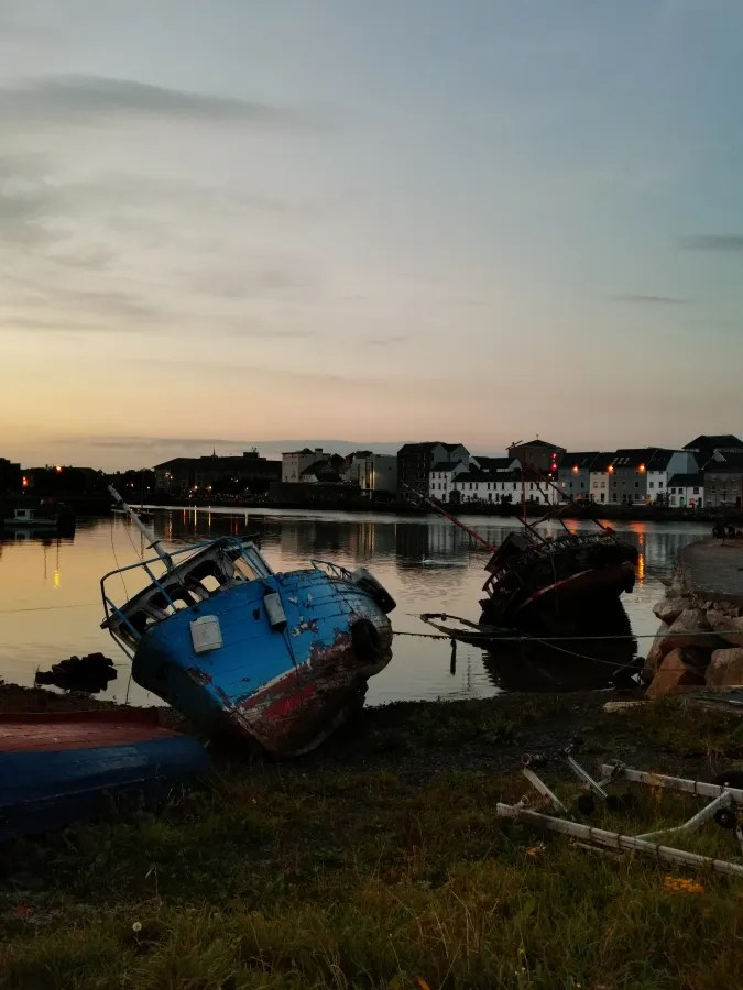 Zwei verlassene Boote am Ufer eines ruhigen Gewässers in Galway während des Sonnenuntergangs, umgeben von sanften Farben des Himmels.