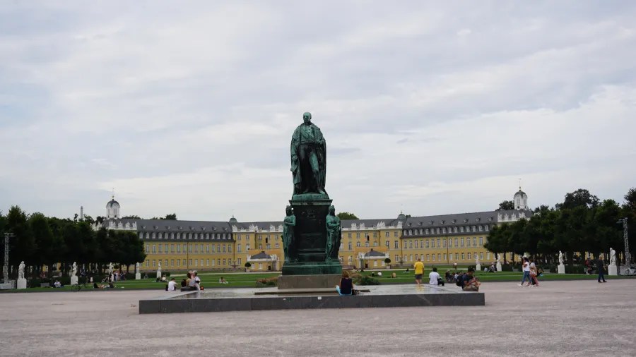 Bronze statue in the foreground with a historical building in the background, situated in a public square with people walking around.