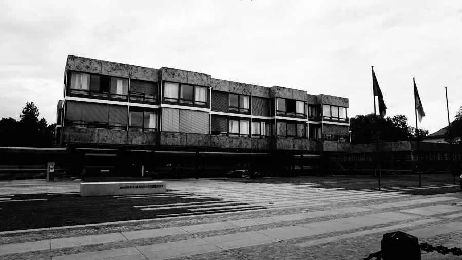 Schwarz-weiß Foto eines modernen, mehrstöckigen Gebäudes mit großen Fenstern, umgeben von einem offenen Platz und Fahnen im Vordergrund.