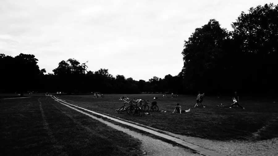 Schwarz-weiß Bild eines weitläufigen Parks mit Menschen, die sich auf der Wiese entspannen und Fahrradfahren. Bäume umgeben den Bereich.