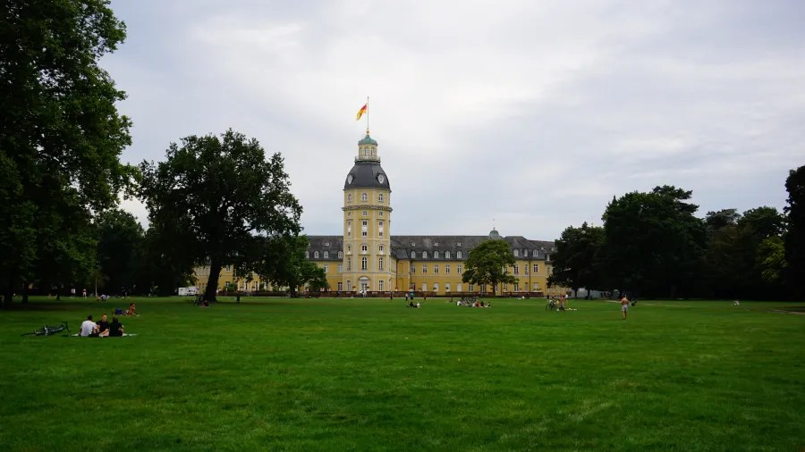 Blick auf das Schloss Karlsruhe mit dem großen, grünen Rasen im Vordergrund, umgeben von Bäumen und Besuchern, die die Umgebung genießen.