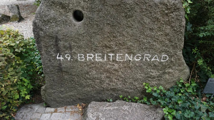 Ein großer Stein mit der Aufschrift '49. BREITENGRAD', umgeben von Pflanzen und grünem Gestrüpp.
