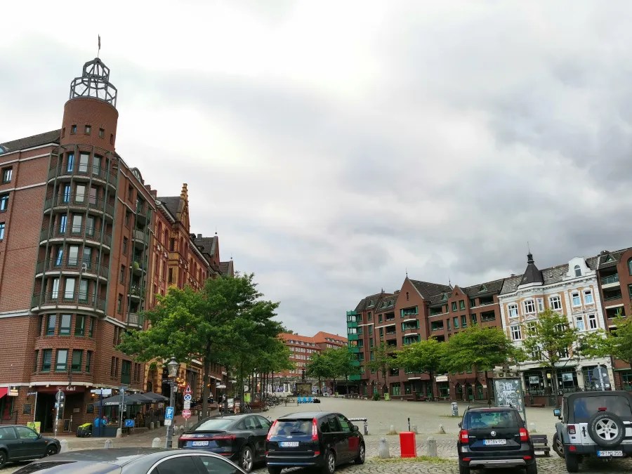 Blick auf einen Platz in Hamburg mit mehrstöckigen, historisch inspirierten Gebäuden und modernen Elementen, umgeben von Bäumen und Fahrzeugen, unter einem bewölkten Himmel.