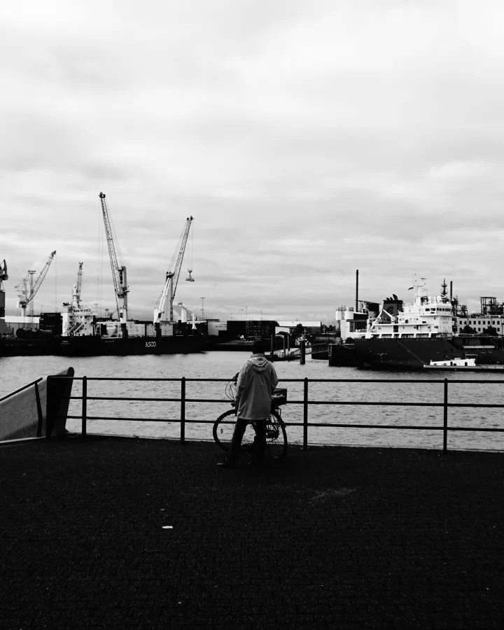 Ein Mensch mit einem Fahrrad steht an einem Hafen und blickt auf die Wasseroberfläche, während im Hintergrund Kräne und Schiffe sichtbar sind. Der Himmel ist bewölkt.