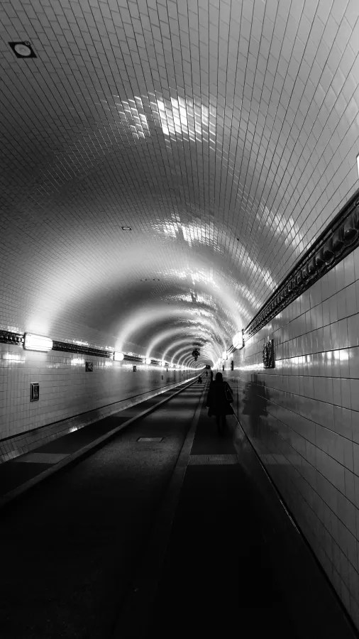 Ein langer Tunnel mit weißen Fliesen, sanft beleuchtet, der den Blick auf den Ausgang in der Ferne freigibt. Im Vordergrund ist eine Person zu sehen, die in Richtung des Ausgangs geht.