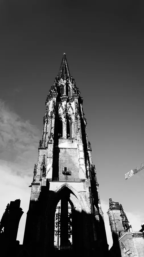 Schwarz-weiß-Bild eines hohen, gotischen Kirchturms mit detaillierten Verzierungen und Schatten im Vordergrund.