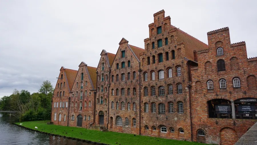 Historische Backsteingebäude am Wasser, die im typischen Hanseatischen Stil erbaut sind, umgeben von Grünflächen und unter einem bewölkten Himmel.