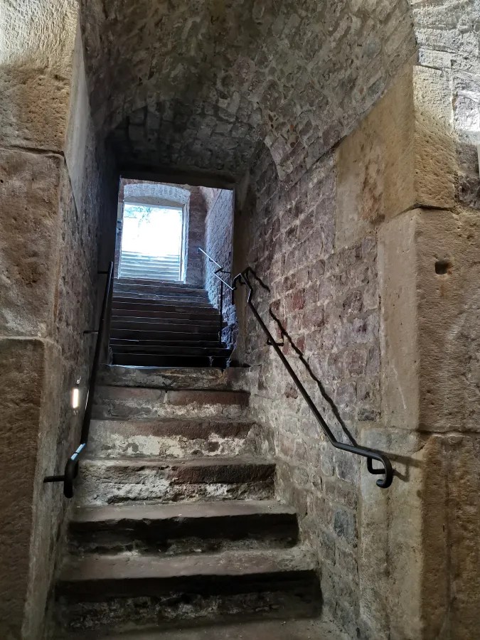 Blick auf eine steinerne Treppe mit Handlauf, die zu einem lichtdurchfluteten oberen Raum führt.