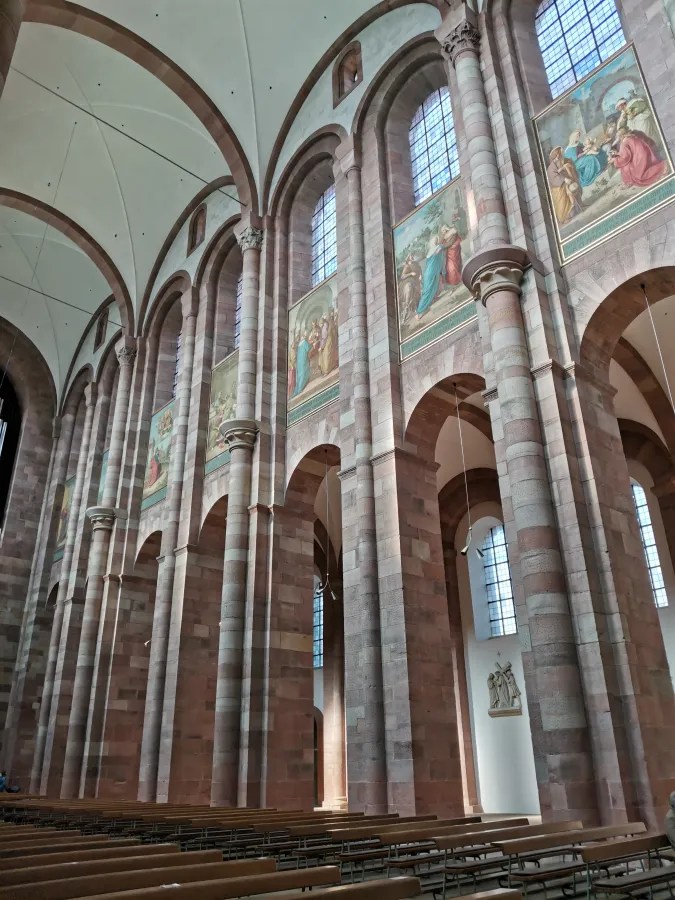 Innenansicht des Speyerer Doms mit hohen Säulen und bunten Fenstern, die historische Wandmalereien zeigen.