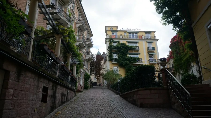Eine steile, gepflasterte Straße in Baden-Baden, flankiert von historischen Gebäuden mit dekorativen Balkonen und üppigem Grün.