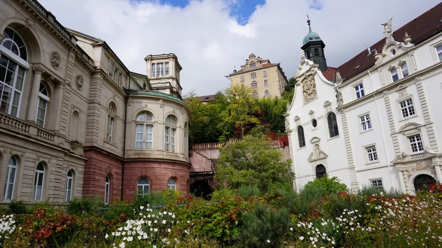 Blick auf historische Gebäude in Baden-Baden, umgeben von üppigem Grün und bunten Blumen unter einem wolkigen Himmel.