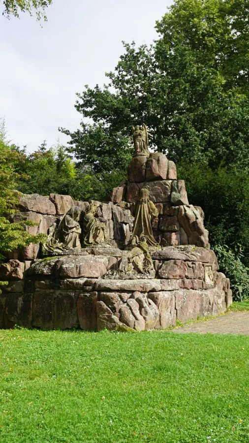 Eine steinerne Skulpturengruppe auf einer Plattform, umgeben von grüner Vegetation und Bäumen, im Freien.