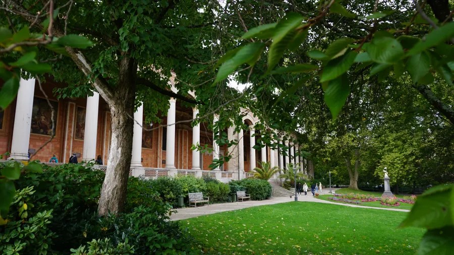 Eine Ansicht des historischen Gebäudes in Baden-Baden, umgeben von grünen Bäumen und einem gepflegten Park mit Bänken und blühenden Pflanzen.