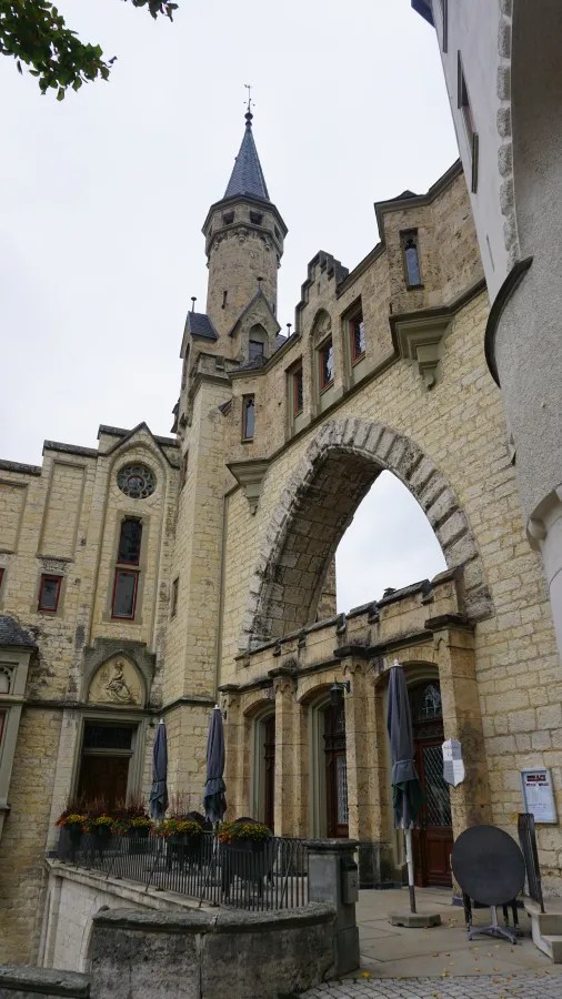 Blick auf ein historisches Gebäude mit einem hohen Turm und steinernen Bogen, umgeben von einem kleinen Biergarten.