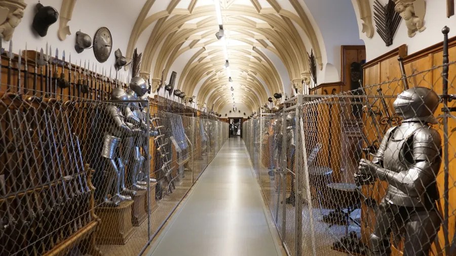 Ein länglicher Korridor mit Rüstungen und Waffen hinter einem Zaun aus Maschendraht, beleuchtet von Deckenlampen.