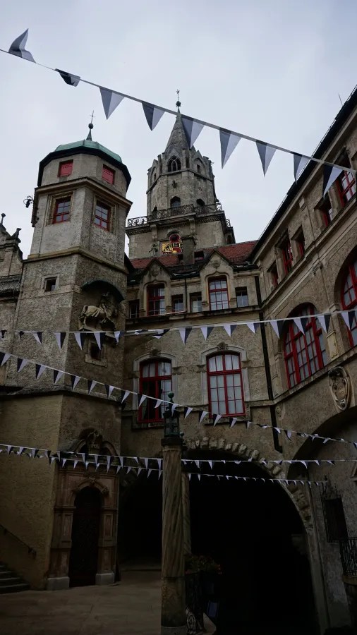 Das Bild zeigt einen Innenhof mit historischen Gebäuden und bunten Wimpeln, die über den Platz gespannt sind. Ein Turm ist im Hintergrund sichtbar, während die Wolken den Himmel bedecken.