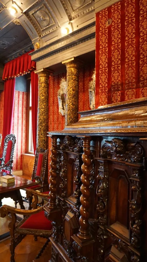 Interior of a richly decorated room featuring ornate wooden furniture, red patterned wallpaper, and tall columns.