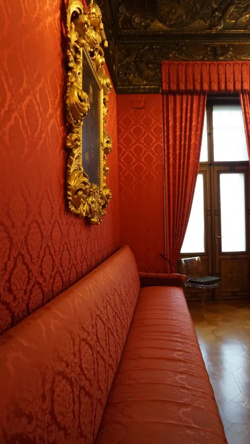 Ein Innenraum mit rotem Wandteppich, einer goldenen Rahmung um ein Gemälde und einem langen roten Sofa, das einen eleganten und historischen Eindruck vermittelt.