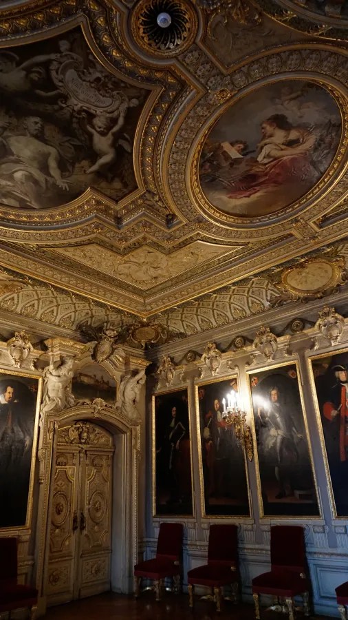 Schöner historischer Raum mit kunstvoll verzierten Wänden und Decke, goldenen Verzierungen und mehreren Portraits an den Wänden.