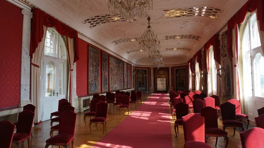 Elegante Innenansicht eines Saals mit roten Wänden, Kristalllüstern und Stuhlreihen auf einem roten Teppich.