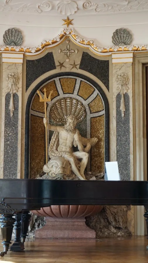 Skulptur eines Meeresgottes auf einer dekorativen Wand mit goldenen und grauen Mosaiken, vor einem schwarzen Klavier.