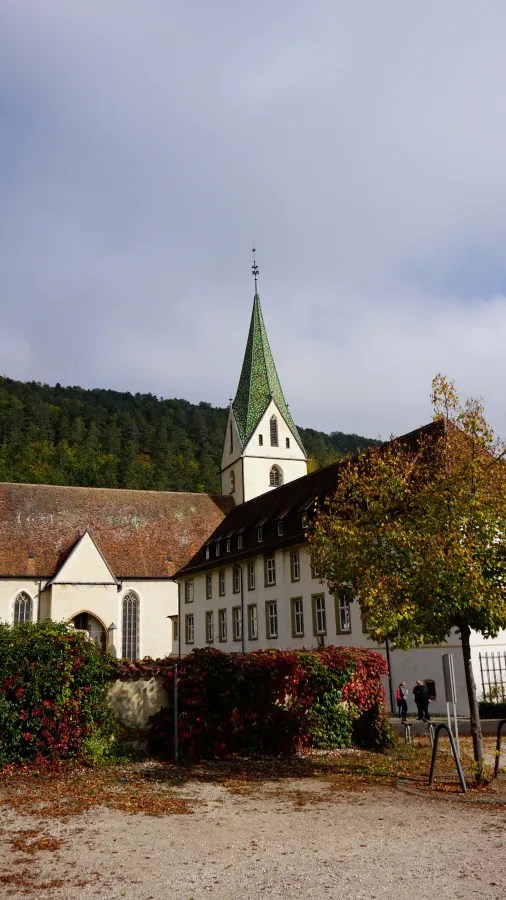 Das Bild zeigt ein älteres Klostergebäude mit einem charakteristischen spitzen Turm und einer grünen Dachdeckung, umgeben von Bäumen und natürlicher Landschaft.