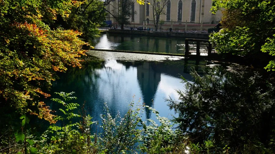 Blick auf den Blautopf in Blaubeuren, umgeben von Bäumen und mit reflektierendem Wasser, während im Hintergrund ein historisches Gebäude sichtbar ist.