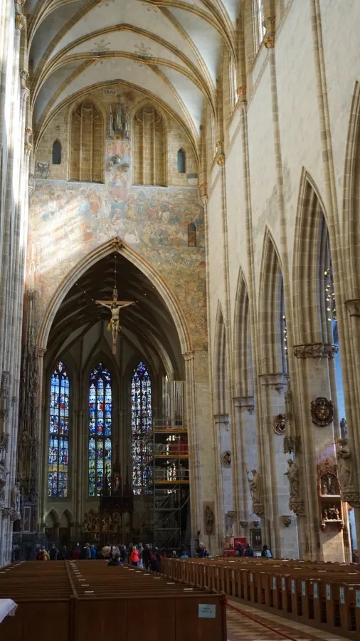 Innenansicht einer Kirche mit hohen Gewölben, bunten Glasfenstern und einem Christus-Kreuz im Bereich des Altars.