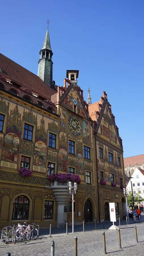 Fassade des historischen Rathauses in Ulm mit bunten Wandmalereien, einem Uhrturm und blühenden Balkonpflanzen.