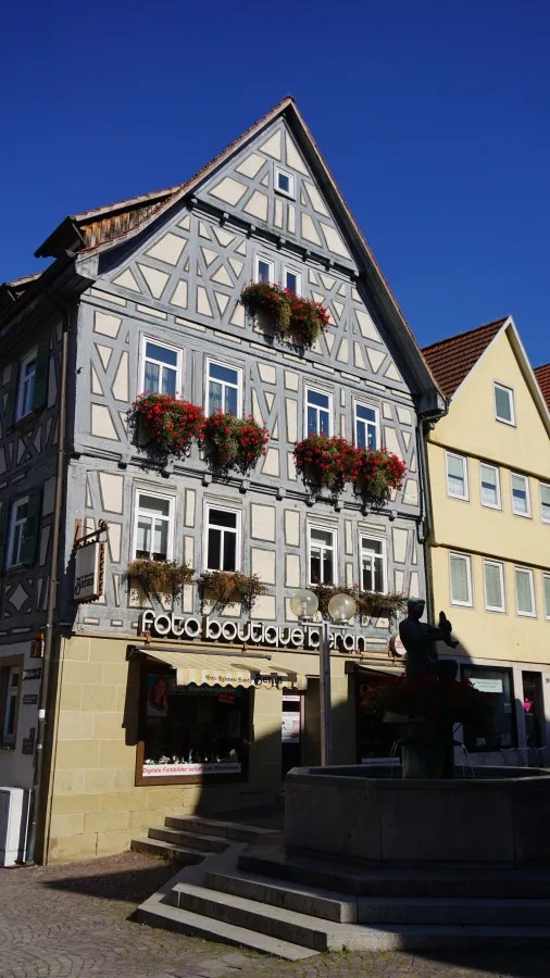 A picturesque building with a timber-framed facade adorned with flower boxes, featuring a photography shop on the ground floor and a fountain in the foreground.
