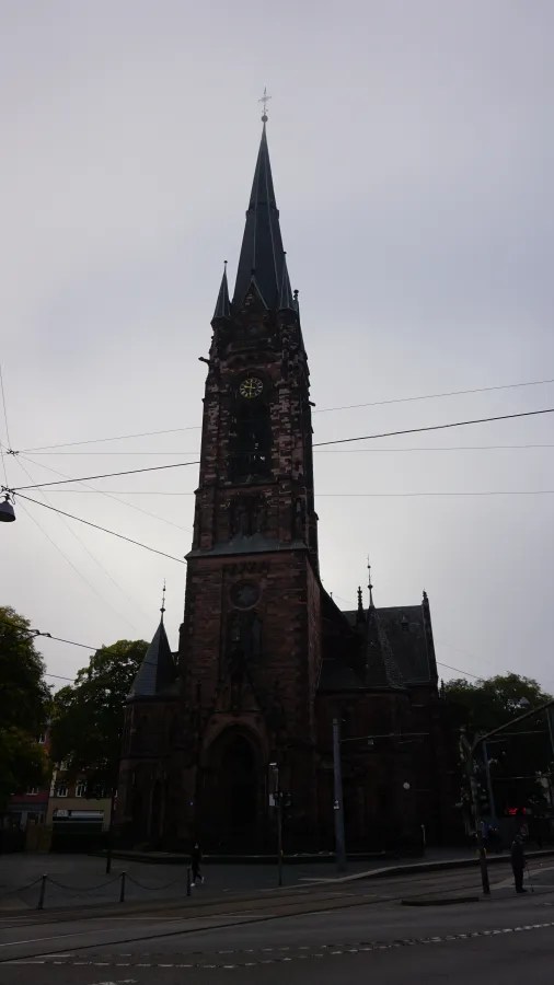 Blick auf eine alte Kirche mit spitzem Turm und Zifferblatt, umgeben von Straßen und Bäumen.