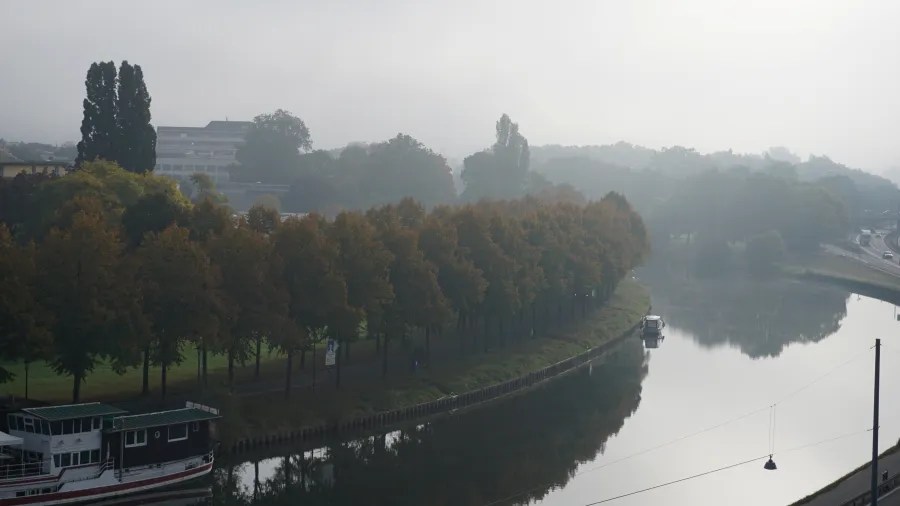 View of a foggy riverbank with autumn trees lining the shore and a boat moored on the water.