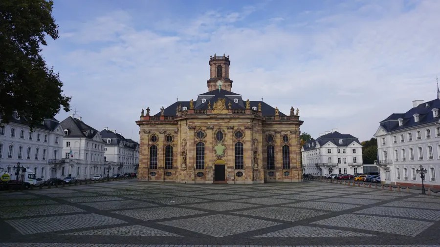 Eine panoramische Ansicht der Ludwigskirche in Saarbrücken, umgeben von klassischen Gebäuden und einem gepflasterten Platz unter einem klaren Himmel.