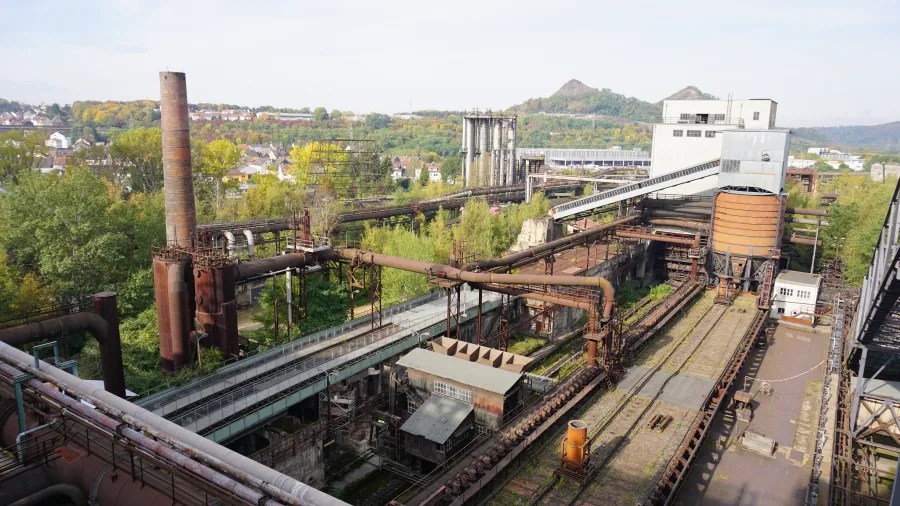Panoramic view of the Völklinger Hütte industrial site showcasing rusted pipes and machinery surrounded by greenery and distant hills.